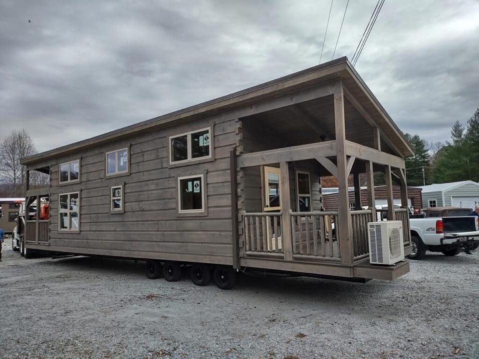 A small, wooden cabin on wheels sits in an outdoor lot. It features multiple windows and a covered porch. The sky is overcast, creating a calm ambiance.