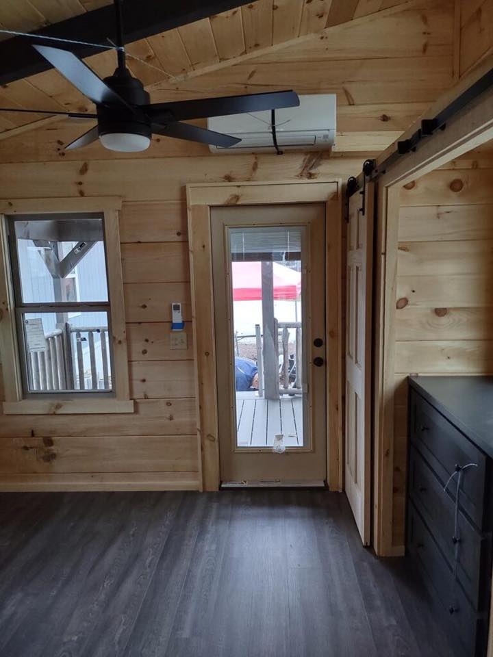 Interior of a wooden cabin with a ceiling fan. A glass door leads outside to a porch. Adjacent, a window adds light, and dark cabinets are on the right.