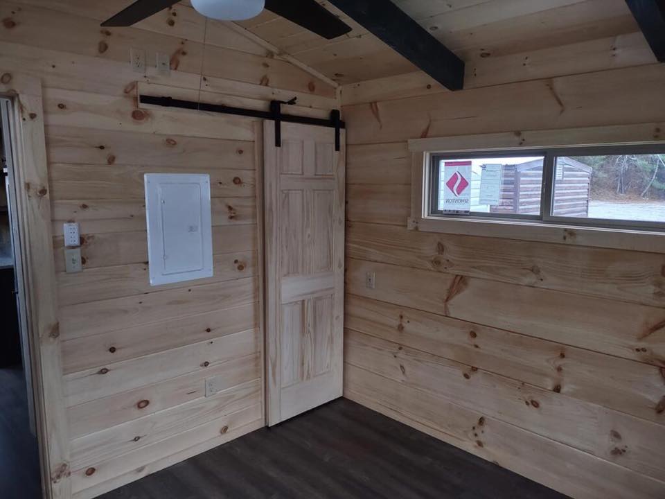 Cozy room with light wood-paneled walls and a dark wood floor. A sliding barn door is centered, and a narrow window lets in soft natural light.