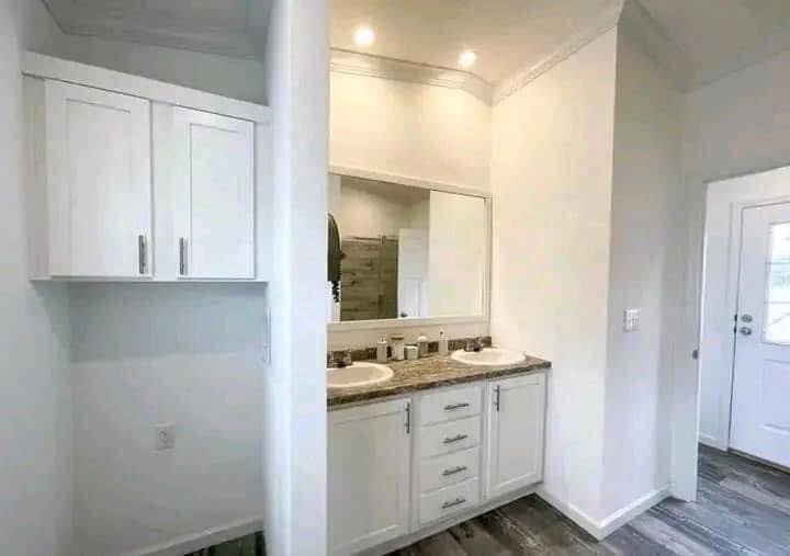Bright bathroom with dual sinks on a granite countertop beneath a large mirror. White cabinets and wood flooring add to the modern, clean feel.