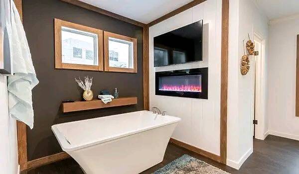 Modern bathroom with a sleek white bathtub, dark accent wall, and mounted electric fireplace. Two framed windows and decorative items create a cozy ambiance.