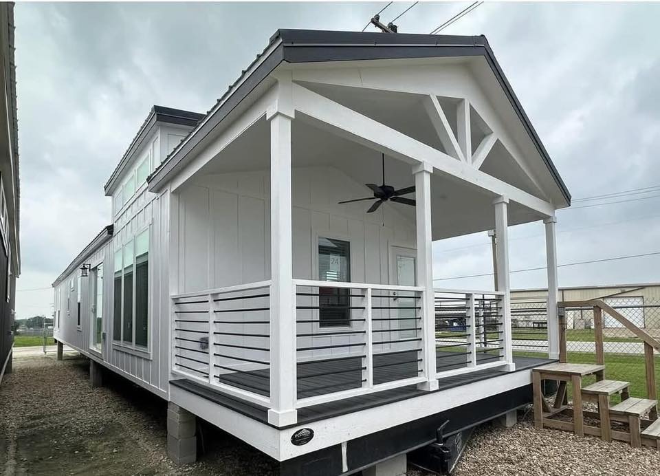 Modern tiny house with white exterior, black roof, and a covered porch featuring a ceiling fan. Wooden stairs lead up; set on a gravel base. Cloudy sky above.