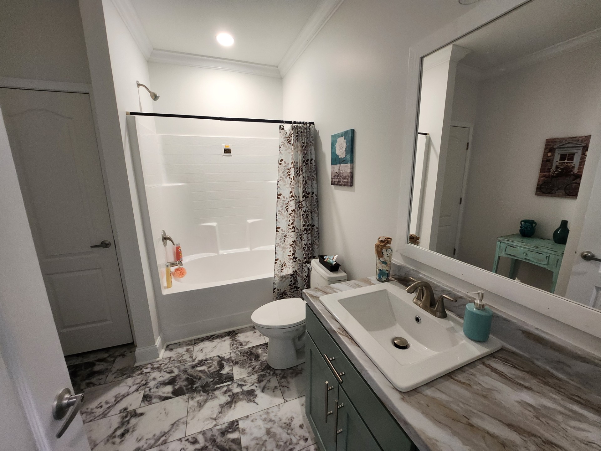 Bright bathroom with marble floor, featuring a white bathtub with a floral curtain. A sink with teal cabinet, matching soap dispenser, and wall art. Cozy and clean atmosphere.