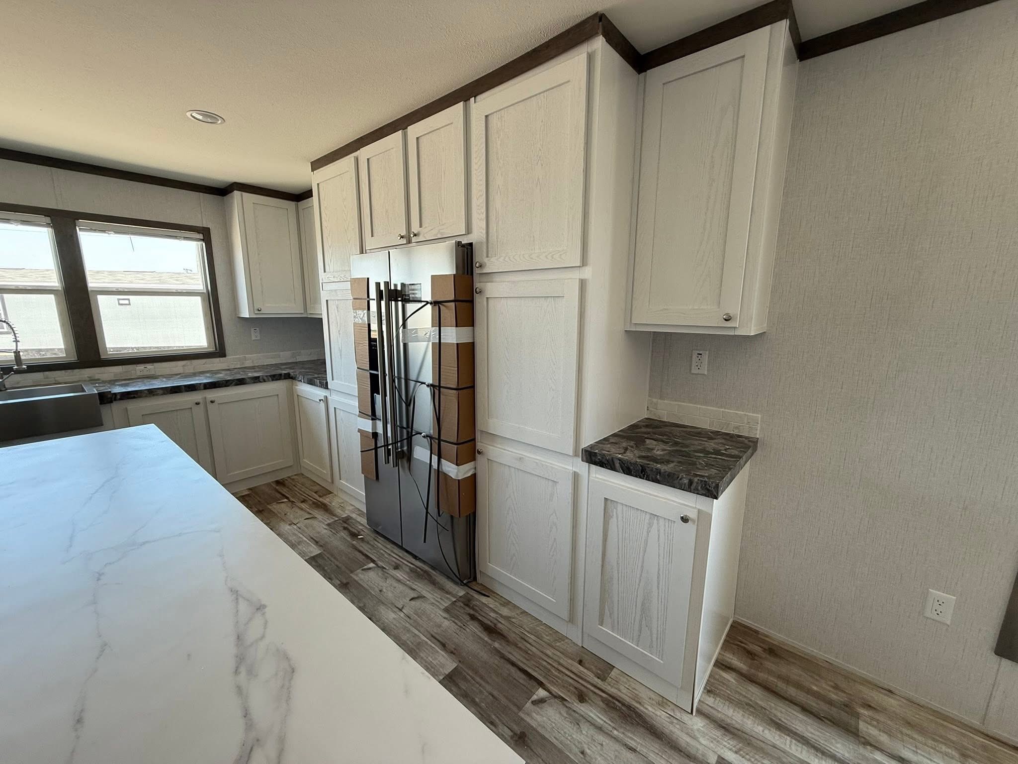 Modern kitchen with white cabinets and marble countertops. A refrigerator is wrapped in protective material. Natural light from a window brightens the space.
