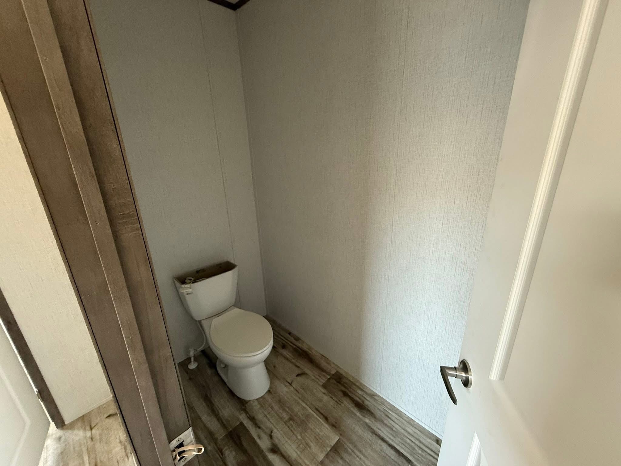Small bathroom with white walls and wooden floor, featuring a toilet near the corner. The open door gives a sense of space and light.