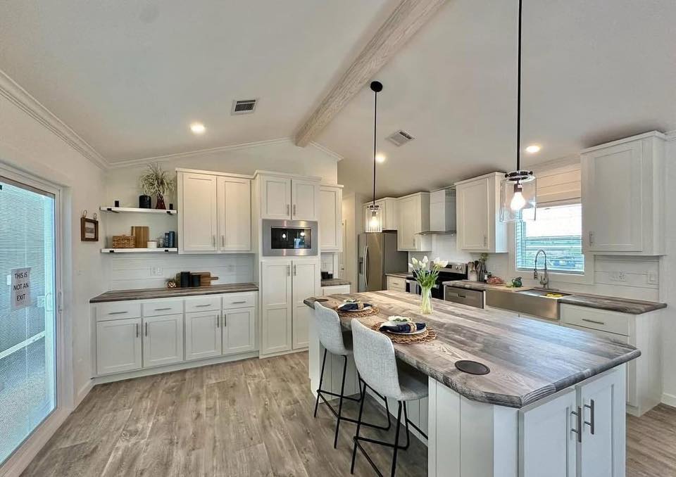Spacious kitchen with white cabinets, wood countertops, and island with stools. Pendant lights hang above island; bright, airy atmosphere.