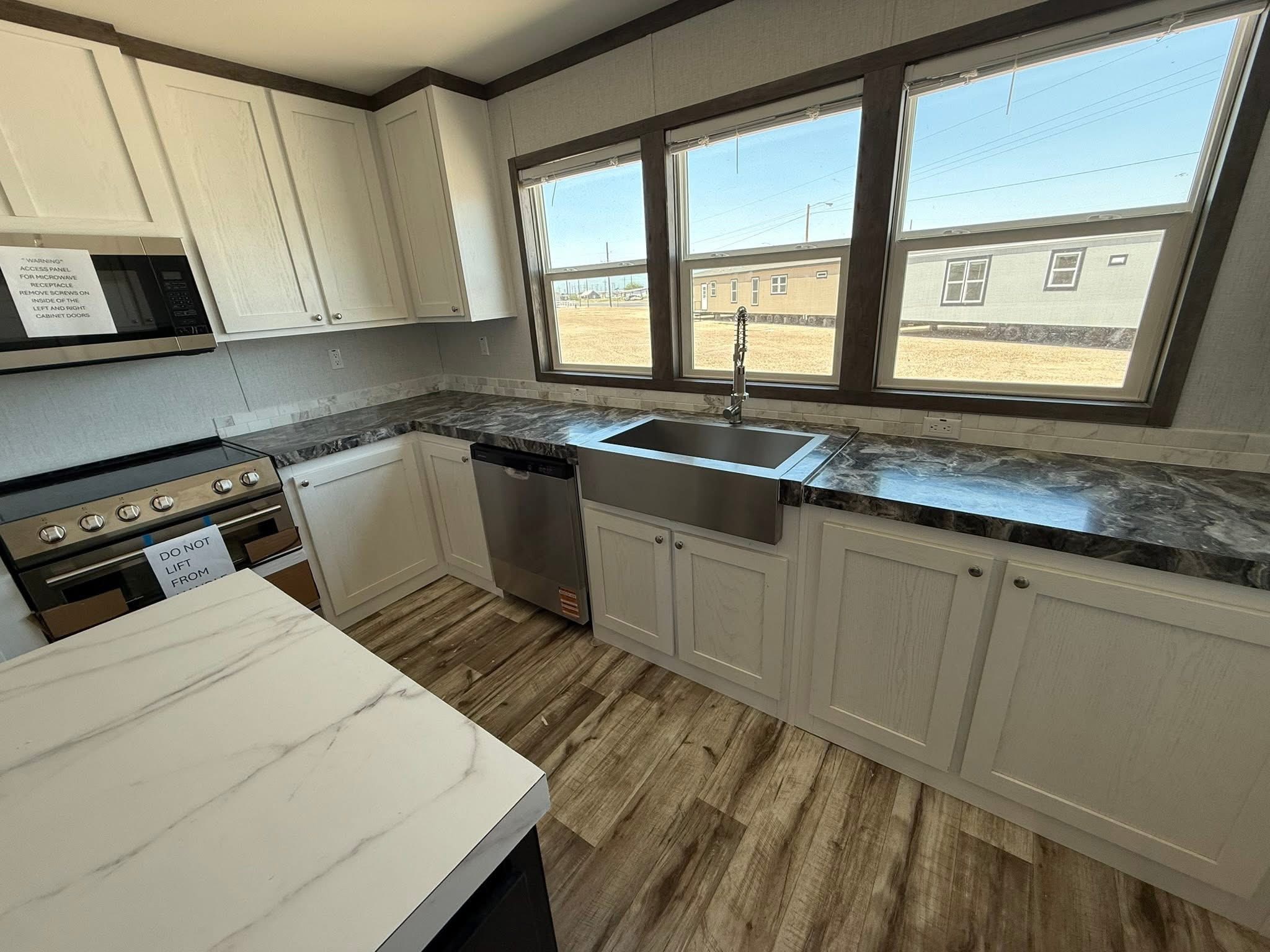Modern kitchen with white cabinets, marble countertops, and stainless steel appliances. Large windows provide natural light, enhancing the sleek design.