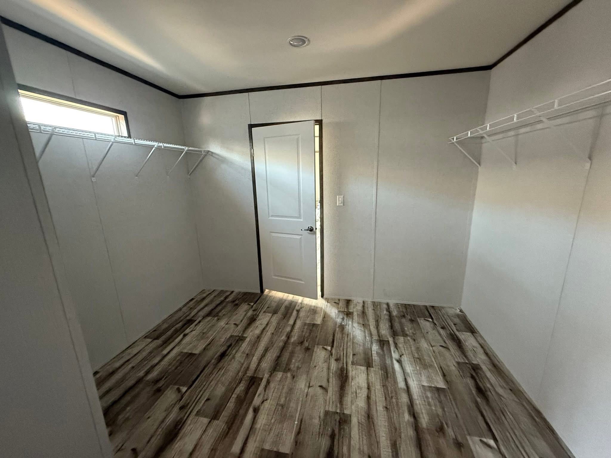 Empty closet room with light gray walls, wooden laminate floors, a single door ajar, and white wire shelves on two sides. Sunlight filters in softly.