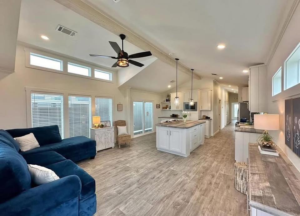 Bright, open-concept living space with a cozy blue sectional, modern kitchen island, light wood flooring, and large windows, creating an airy, inviting atmosphere.