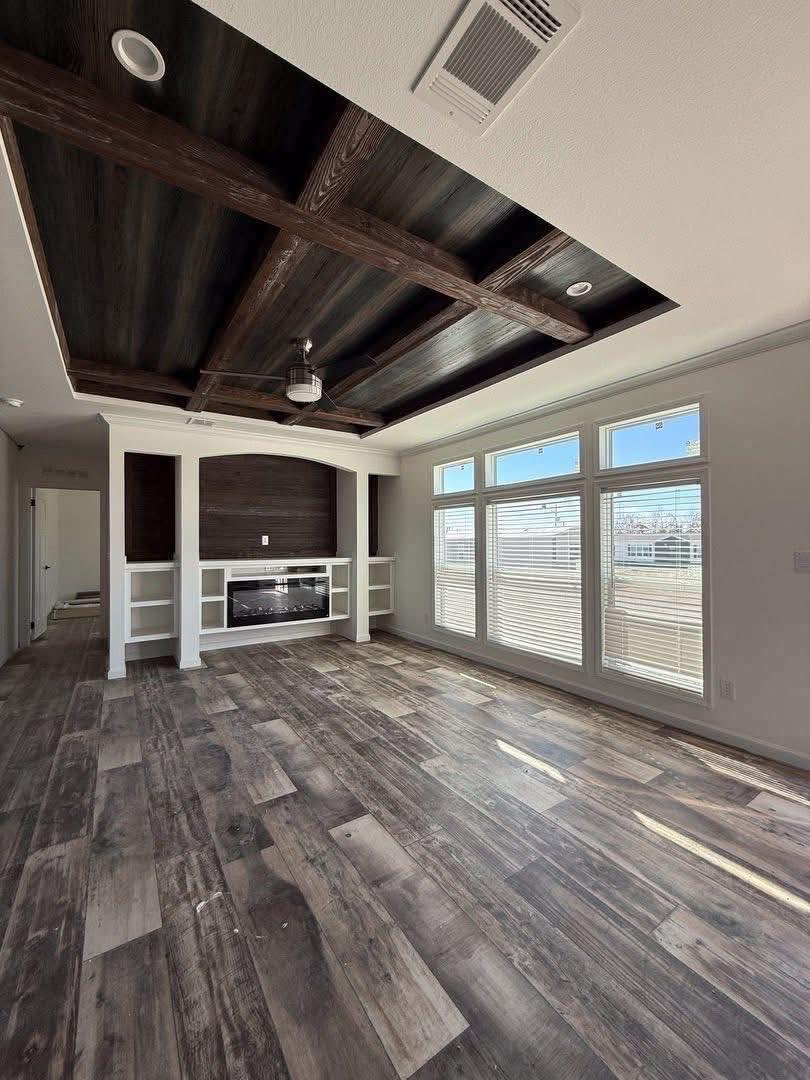 Spacious room with dark wood beamed ceiling and floor, large windows on right letting in sunlight, and a built-in fireplace with shelves on back wall. Cozy, inviting atmosphere.