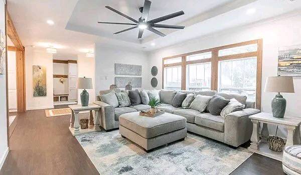 Modern living room with a large gray sectional sofa, a central ottoman, and green accent lamps. Bright windows, cozy ambiance, and elegant decor.