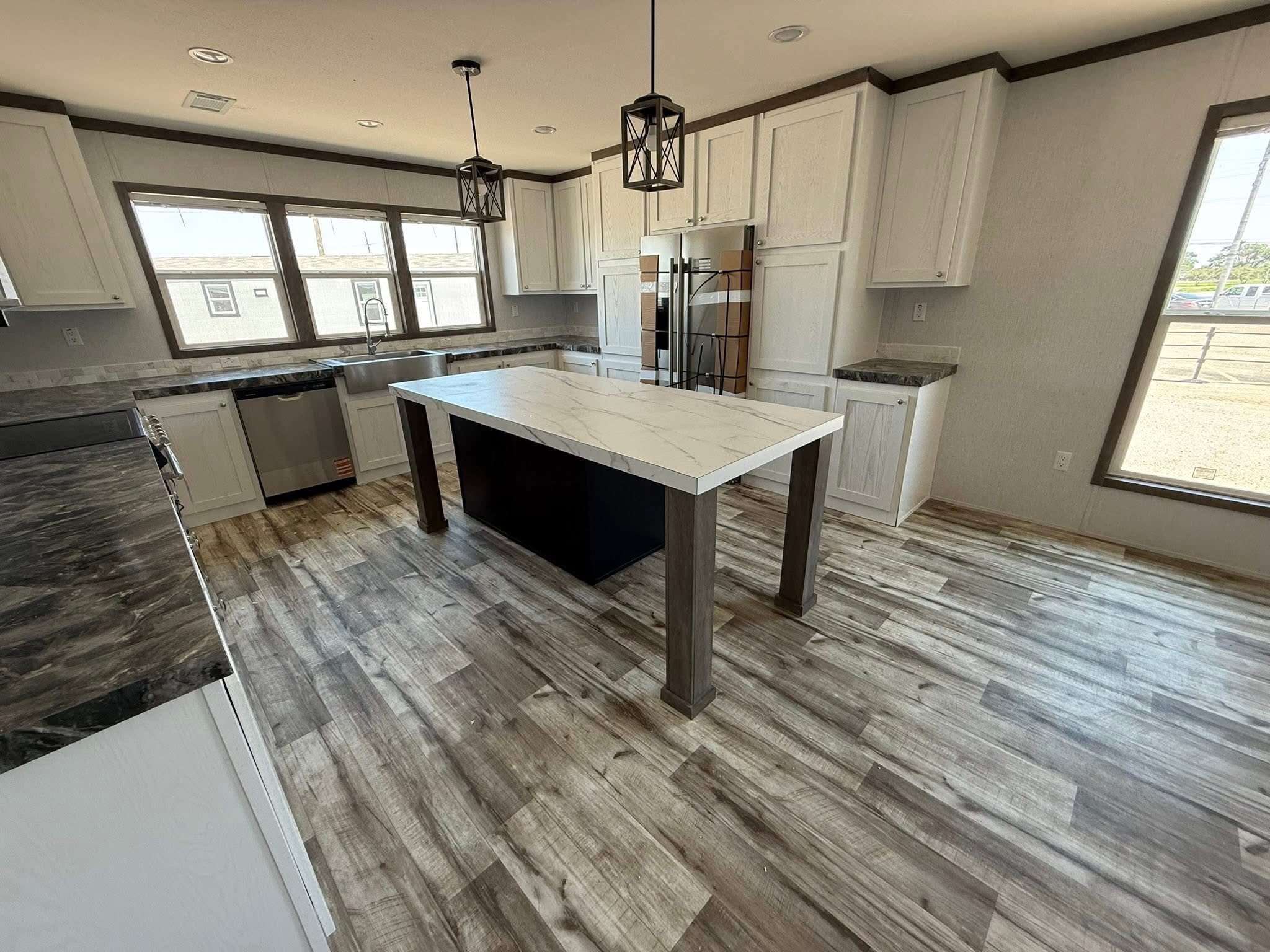 Spacious modern kitchen with wooden flooring, marble island, pendant lights, and light cabinetry. Sunlight streams through large windows, creating a welcoming atmosphere.