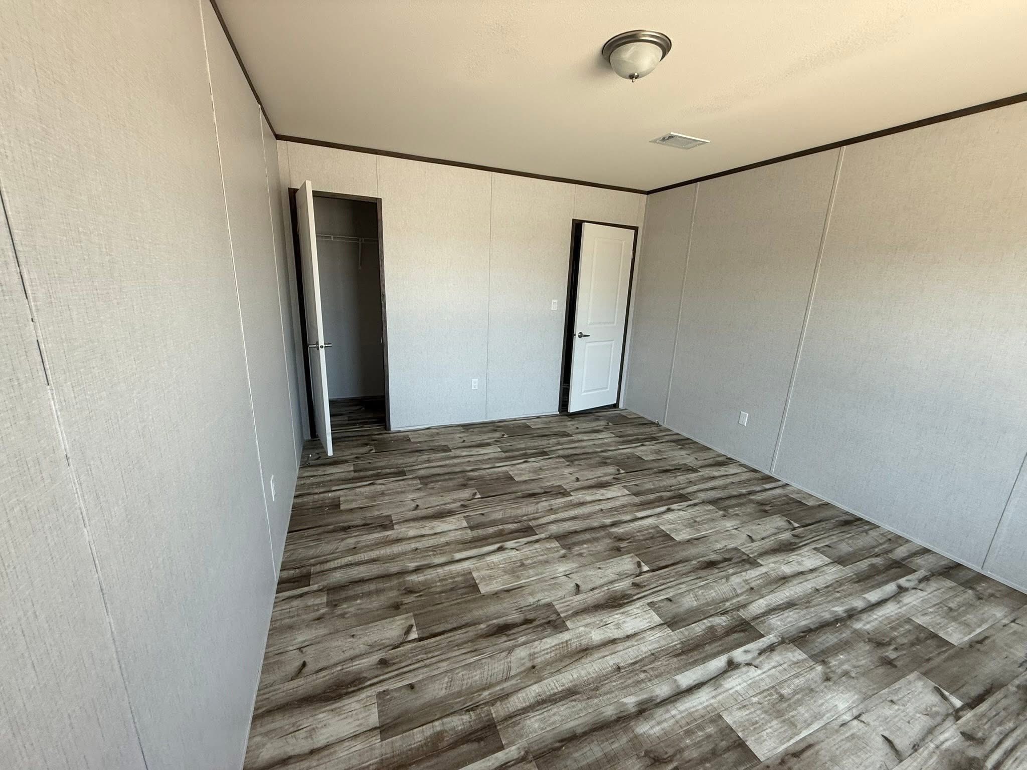 Empty room with neutral-colored walls and wood-patterned vinyl flooring. A closed white door and open closet door are visible. The space feels clean and minimalistic.