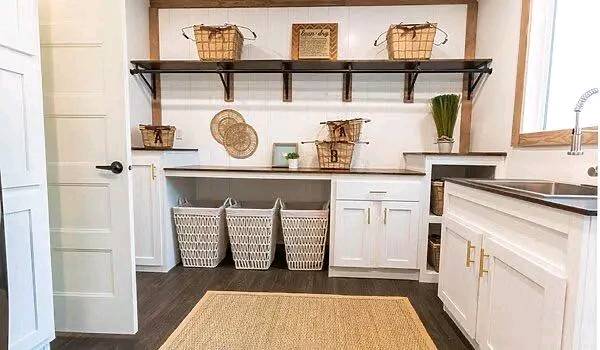 Bright and organized laundry room featuring white cabinets, wicker baskets on shelves, and three laundry hampers below. A woven rug covers the dark wood floor.