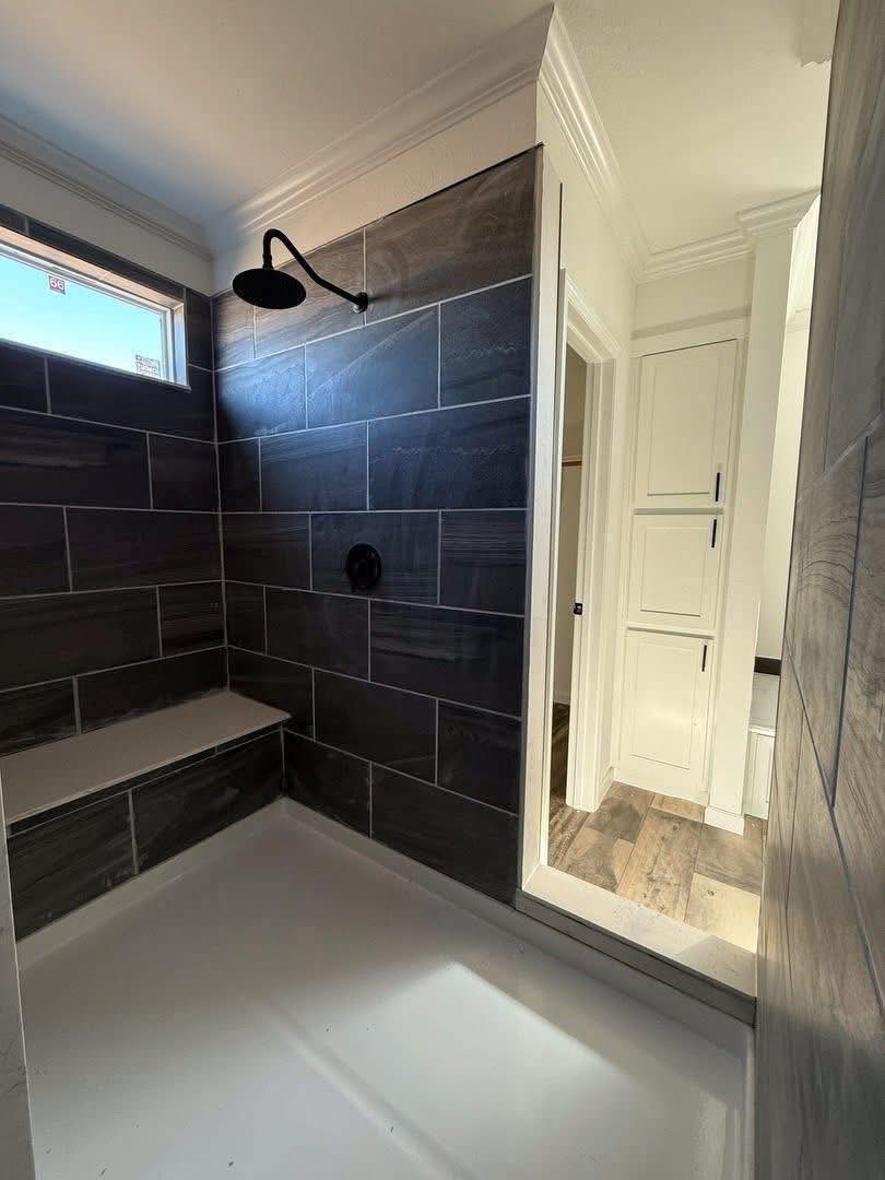 Modern bathroom shower, featuring dark gray large tiles, a black rain showerhead, a white bench, and a narrow frosted window above, conveying a sleek, clean aesthetic.