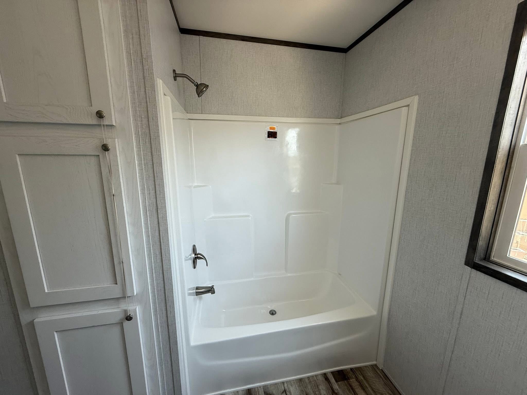 A modern bathroom features a white shower-tub combo with chrome fixtures, adjacent to white cabinets. Natural light streams through a window, enhancing the serene ambiance.