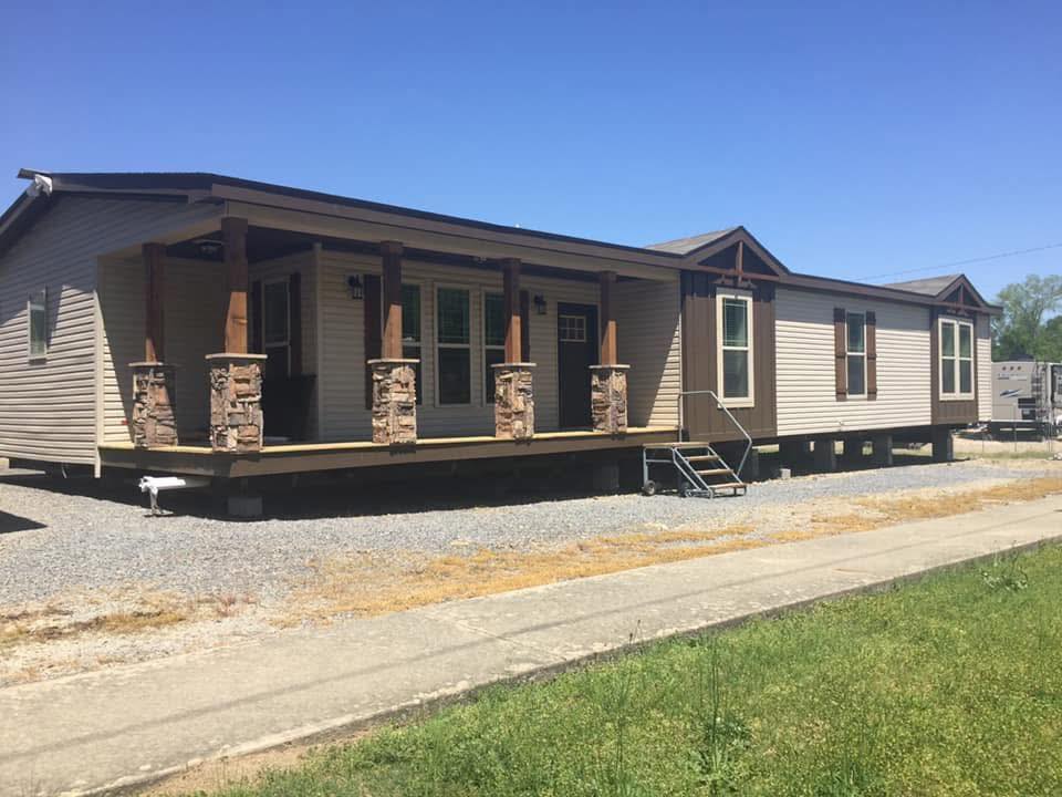 Single-story modular home with beige siding and brown accents. Features a covered front porch with stone pillars and steps, set on gravel. Clear blue sky.
