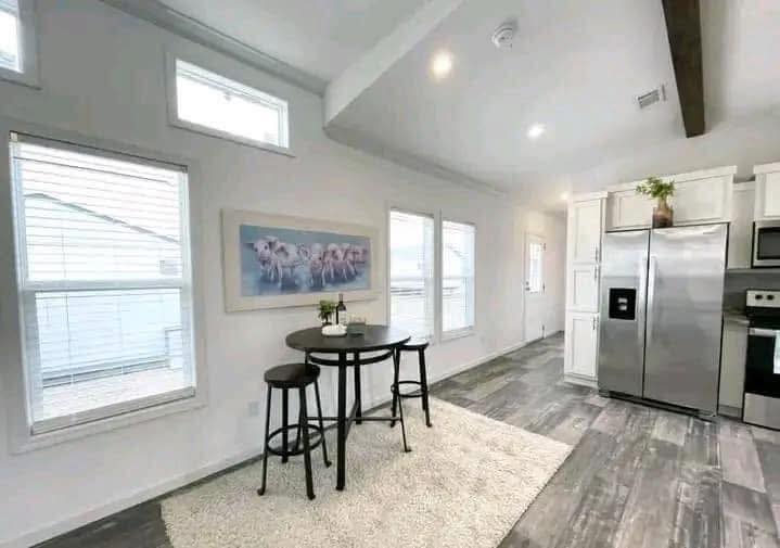Modern kitchen and dining area with white walls, wooden flooring, and natural light. A round table, stools, abstract art, and stainless steel appliances enhance the cozy ambiance.