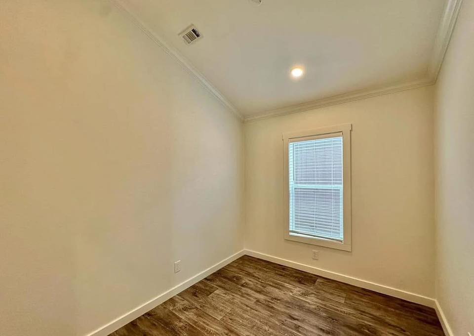 Small, empty room with beige walls and a high ceiling. A single window with blinds lets in light, highlighting the wood-patterned floor. The space feels clean and minimal.