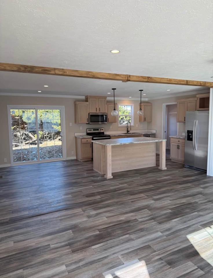 Bright modern kitchen featuring light wood cabinets and island, stainless steel appliances, pendant lights, and sliding glass doors with a view outside.