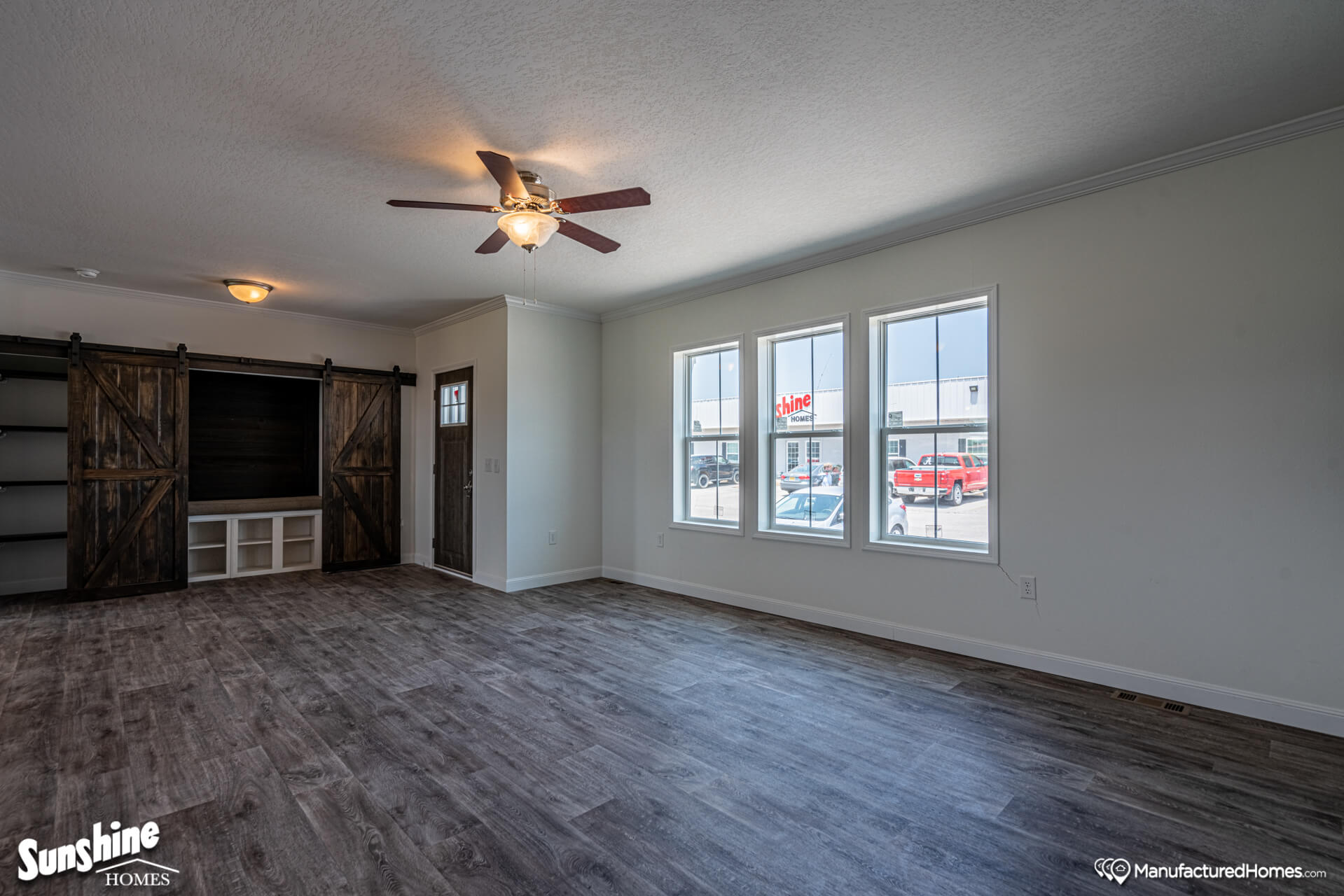 Spacious living room with wood flooring, three large windows, a ceiling fan, and built-in shelves with rustic sliding barn doors. Bright and modern vibe.