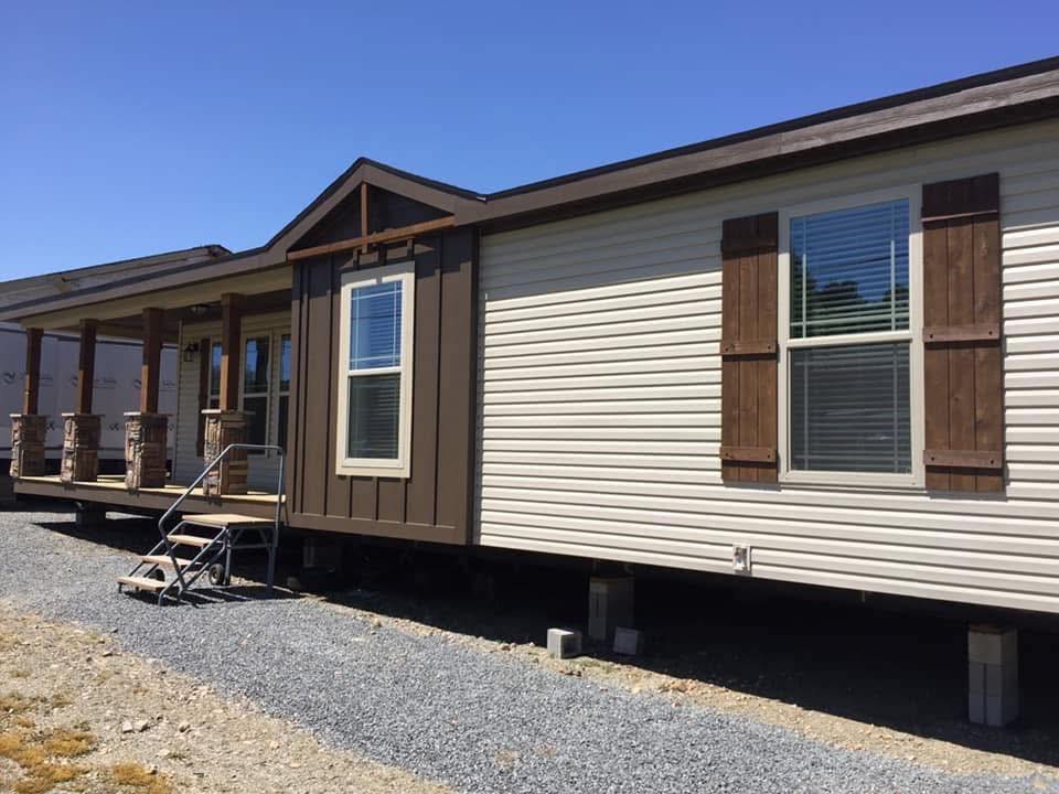 A manufactured home with cream siding and brown shutters sits elevated on concrete blocks over a gravel surface, with a small porch and clear blue sky.