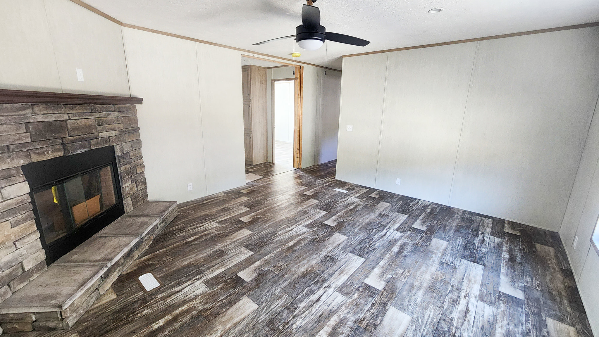 Empty room with rustic wood flooring and light-colored walls. Features a stone fireplace, ceiling fan, and doorway leading to another room. Cozy atmosphere.