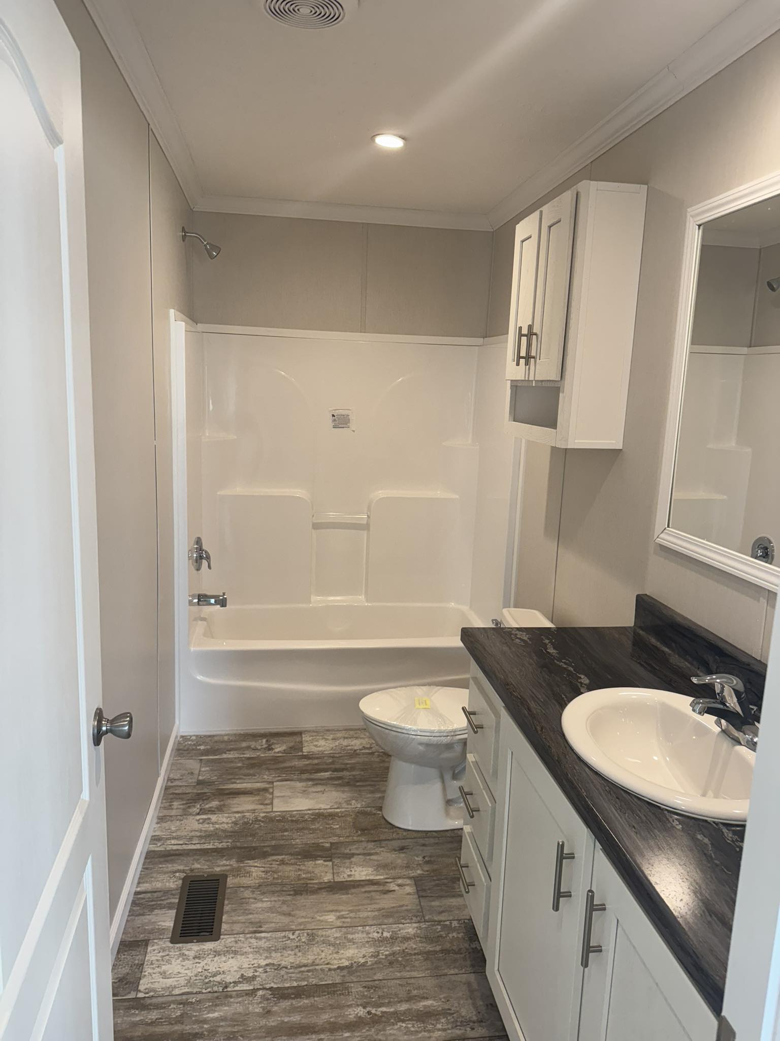 Compact bathroom with white walls features a bathtub-shower combo, a toilet, and a dark countertop vanity. Gray wood-patterned flooring adds a modern touch.