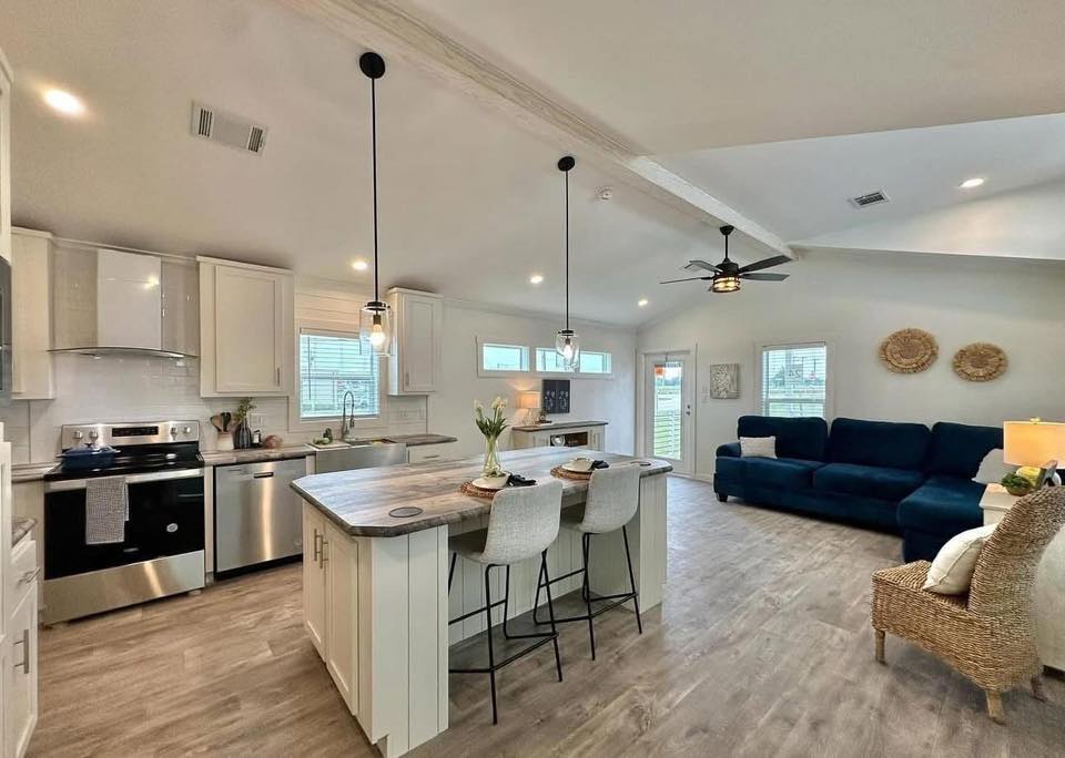 Modern open-plan kitchen and living room with light wood flooring. Features a central island with barstools, pendant lights, and a navy sectional sofa.