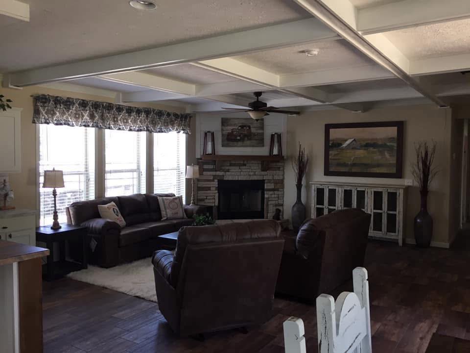 Cozy living room with brown leather sofas, rustic stone fireplace, large windows with curtains, wooden floor, a painting, and a ceiling fan.