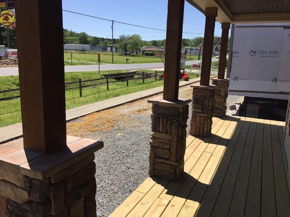 Wooden porch with stone columns under a sunny sky, overlooking a rural road and grassy field. Calm and inviting atmosphere.