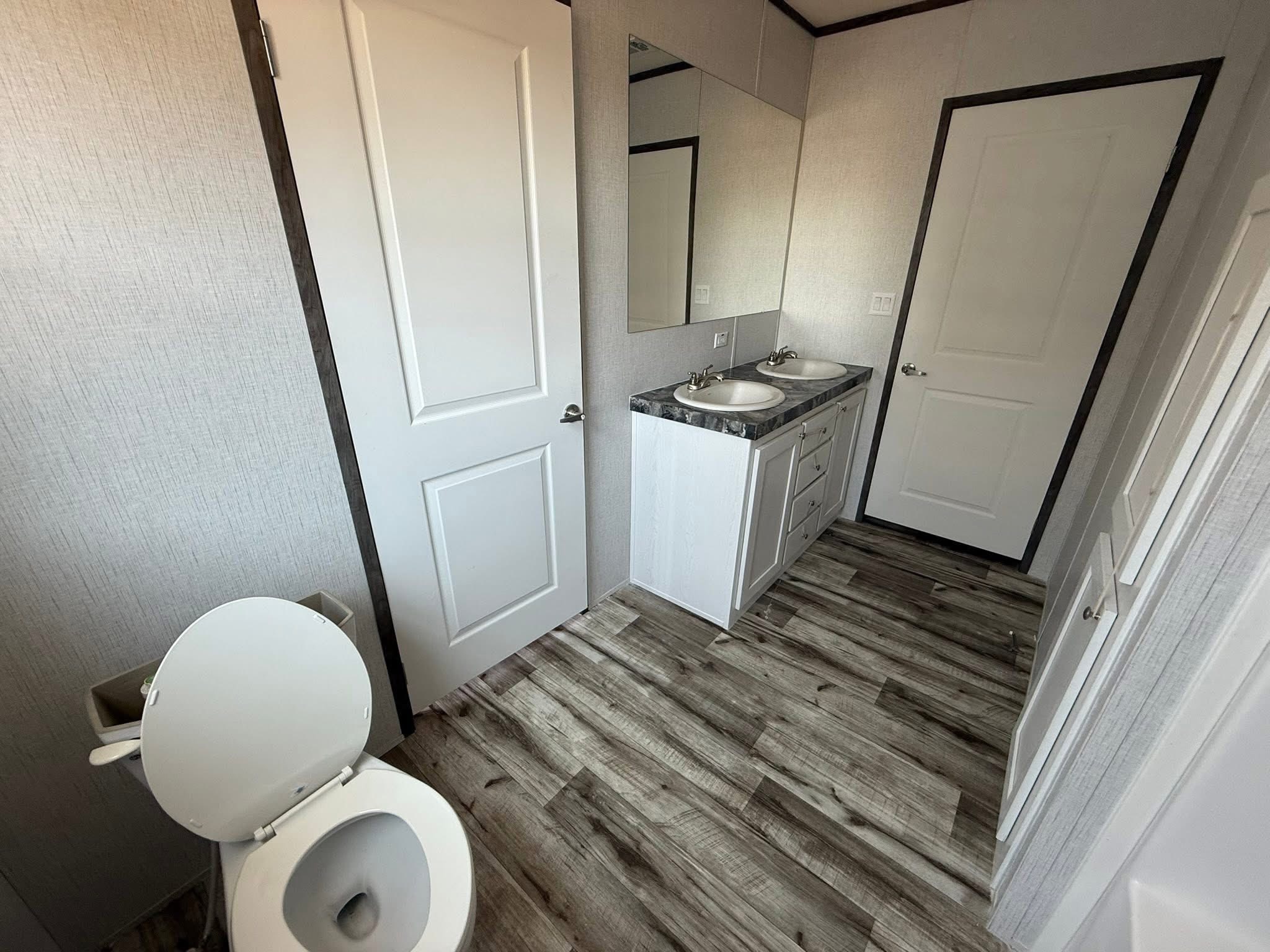 Modern bathroom with light wood flooring, featuring a toilet with an open lid, a vanity with dual sinks, a large mirror, and two closed white doors.
