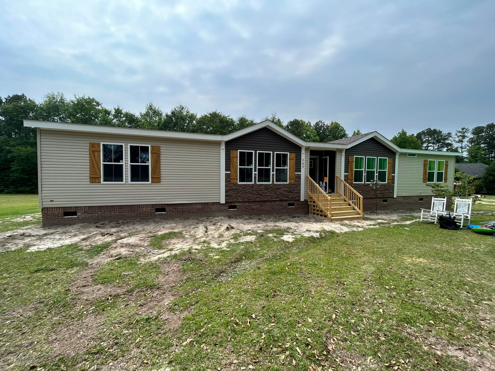 A single-story manufactured home with beige siding and wooden shutters stands on a grassy lawn. The front steps lead to a porch, conveying a peaceful, rural setting.