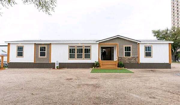 A modern, single-story modular home with a brown and white exterior. It features multiple windows and a central entrance with wooden steps, set in a spacious lot.