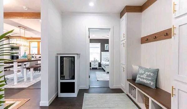 Hallway with white walls and dark wood flooring. A bench with a cushion reads "We are all dreamers." The hallway leads to a bright bedroom and an open kitchen.