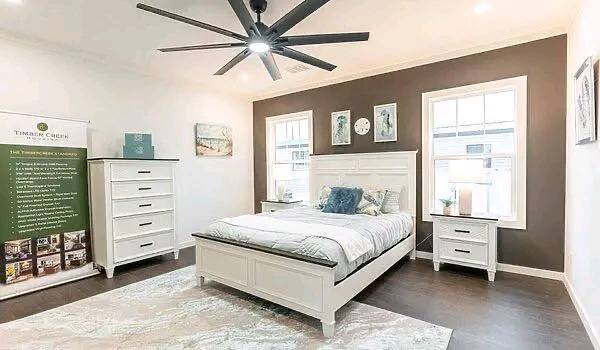 A cozy bedroom features a white bed, dresser, and nightstand with a dark accent wall. Soft lighting, a large ceiling fan, and serene decor create a calming atmosphere.