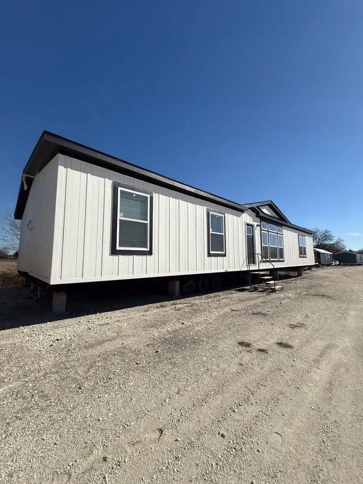 Single-story mobile home with light siding and dark trim, set on a dirt lot under a clear blue sky. Stairs lead to the entrance.