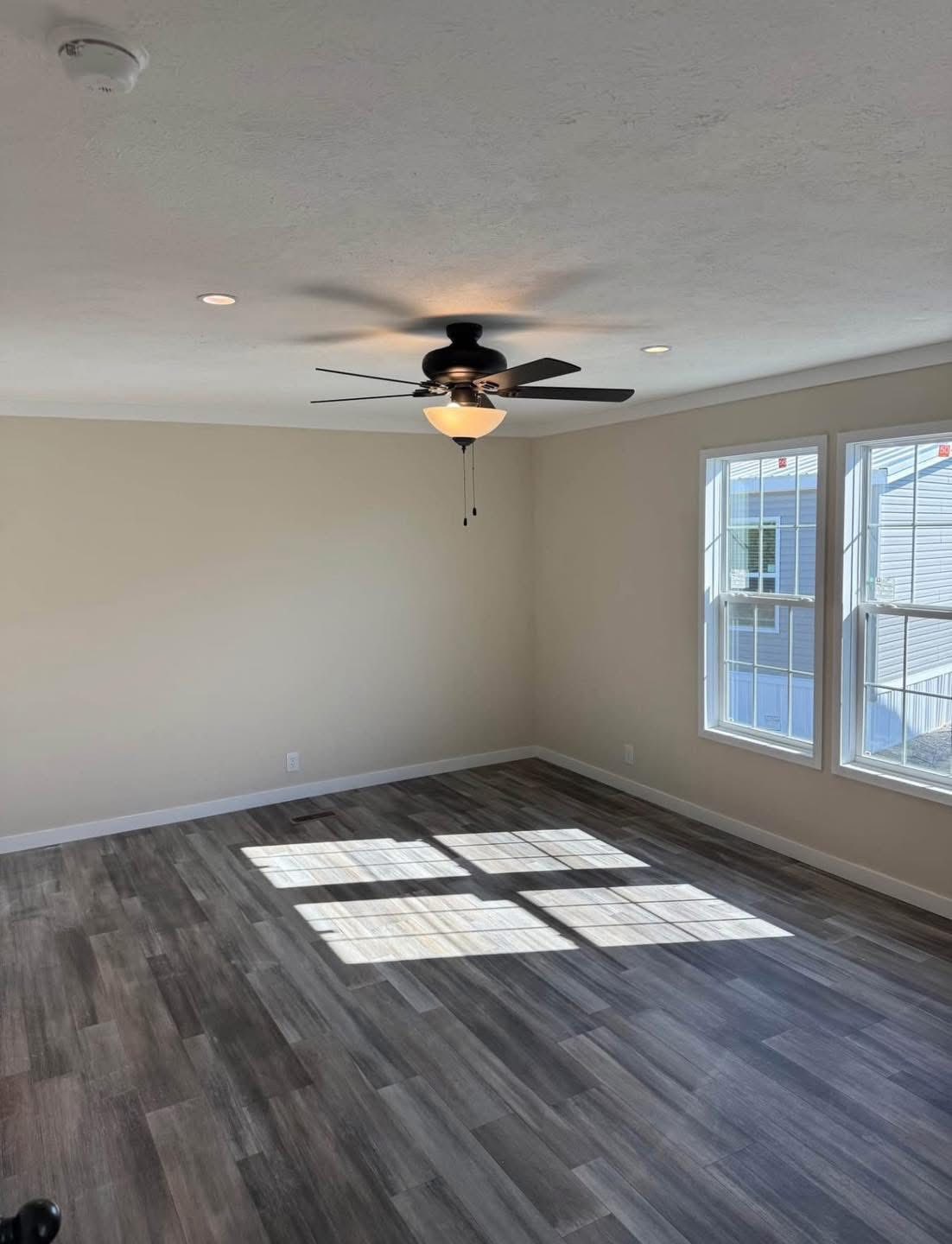 Spacious, sunlit room with beige walls and dark wood laminate flooring. A ceiling fan hangs above, and large windows cast bright light patches on the floor.