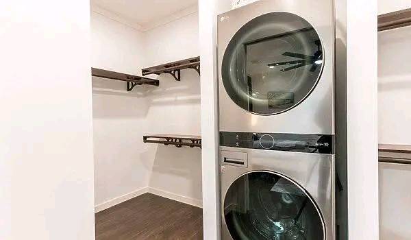 A modern laundry room with a sleek, stainless steel stacked washer and dryer. Empty wooden shelves line the white walls, creating a minimalist feel.