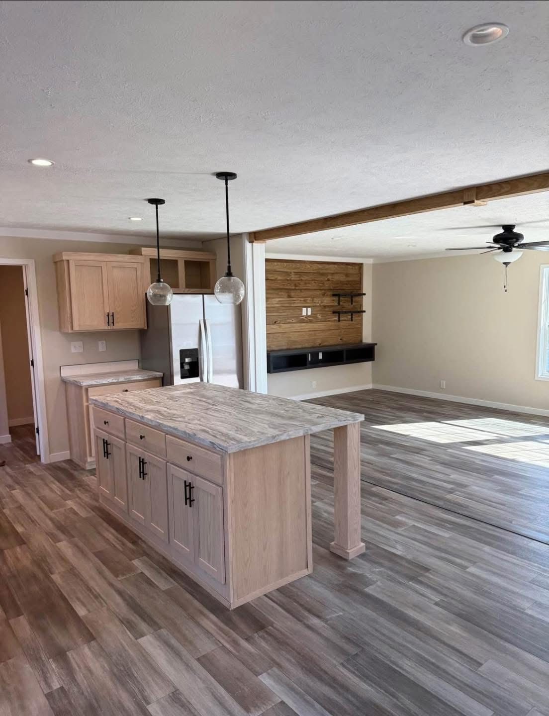 Modern kitchen and living area with light wood flooring. Features a central island, stainless steel fridge, ceiling fan, and accented wood wall. Bright and airy ambiance.