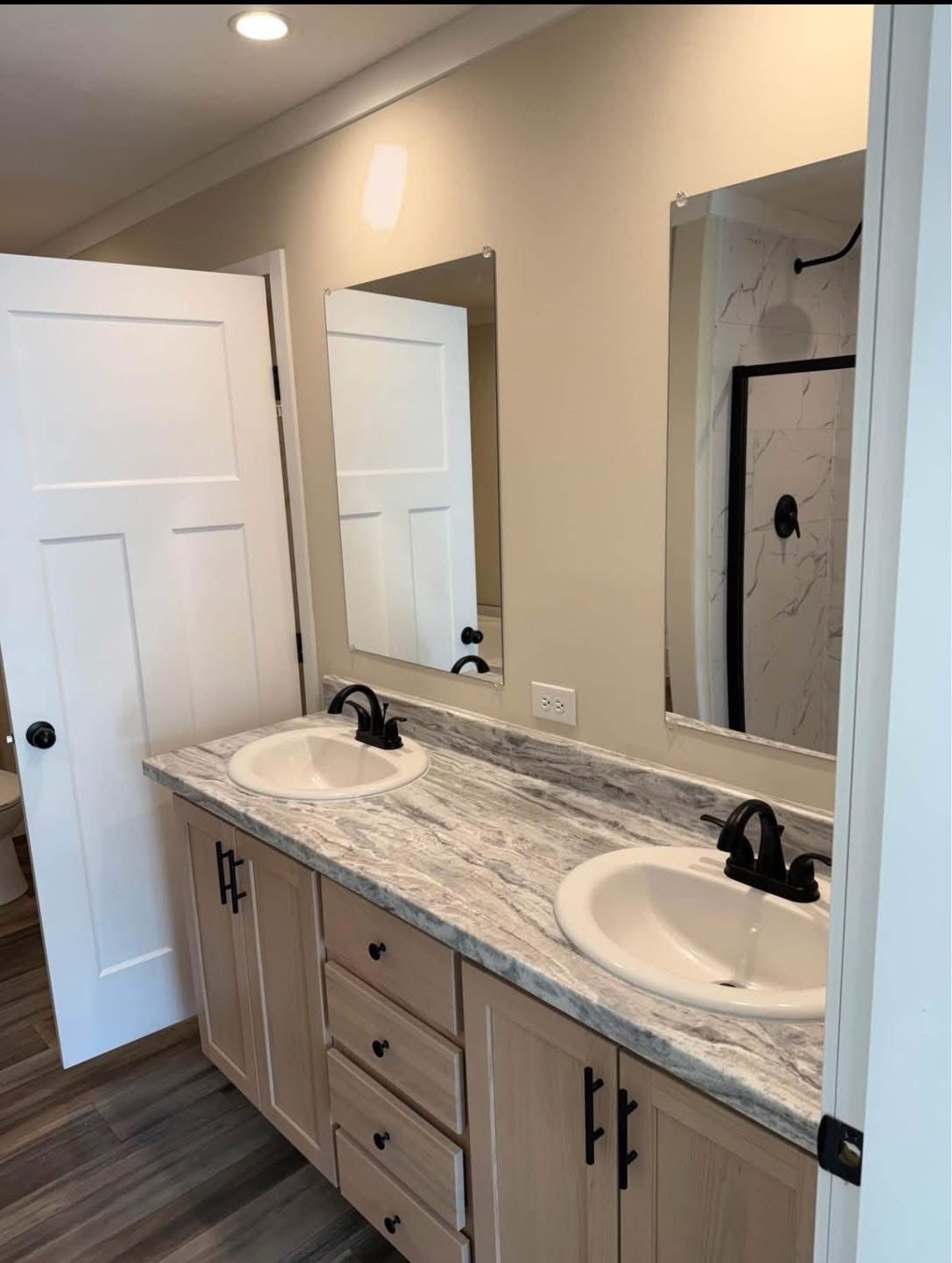 Modern bathroom with light wood double vanity and marble countertop. Two white sinks have black faucets. Beige walls and large mirrors reflect a shower.