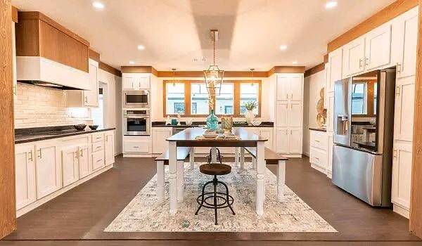 A spacious, modern kitchen with white cabinetry, a central wood dining table, and stainless steel appliances. Warm lighting creates a cozy atmosphere.