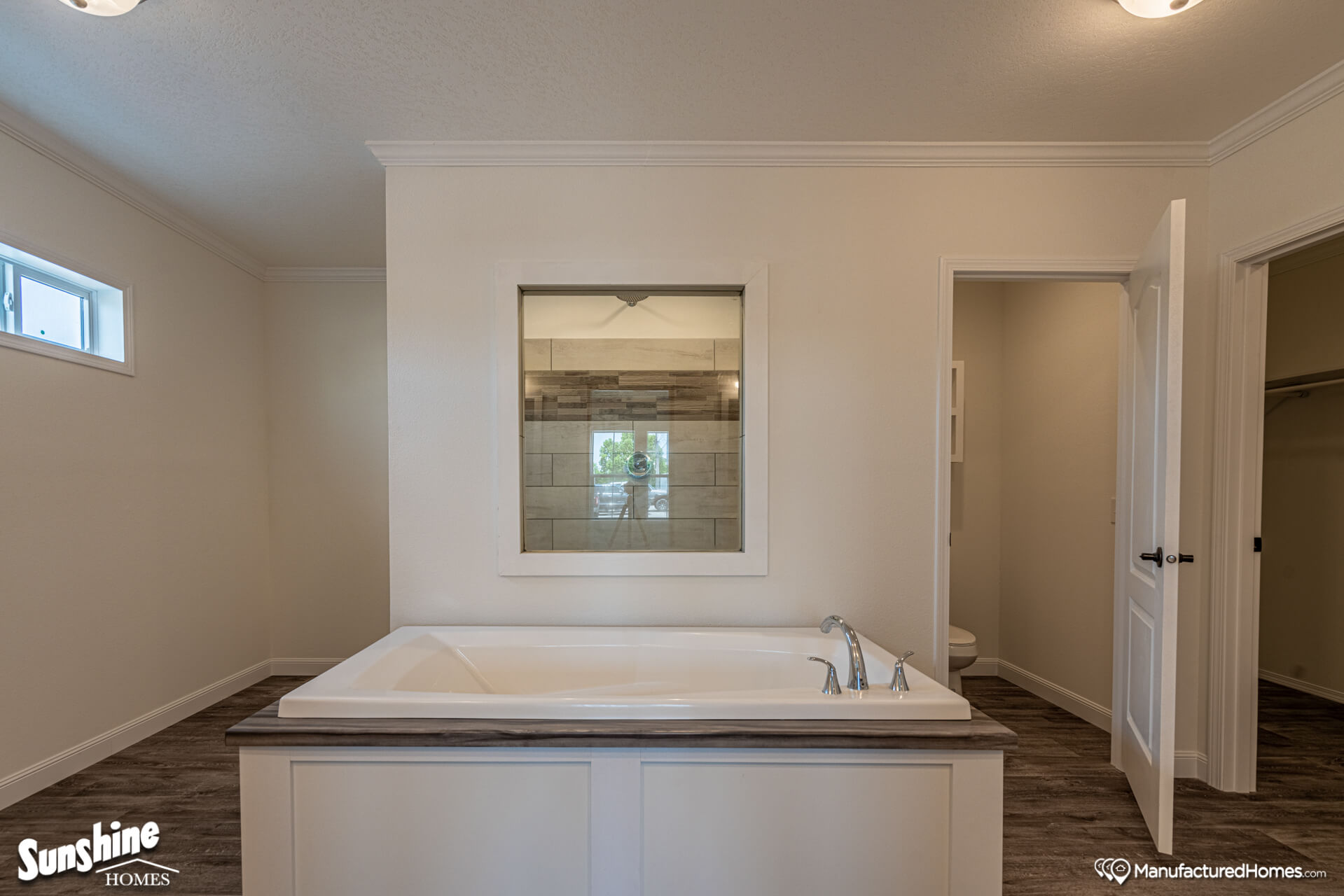 A modern bathroom featuring a central white tub with sleek silver faucets, set against a neutral wall. A window in the background adds natural light.