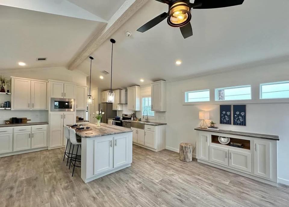Spacious modern kitchen with white cabinets, island with pendant lights, and wood flooring. Ceiling fan, natural light, and serene decor create a cozy ambiance.