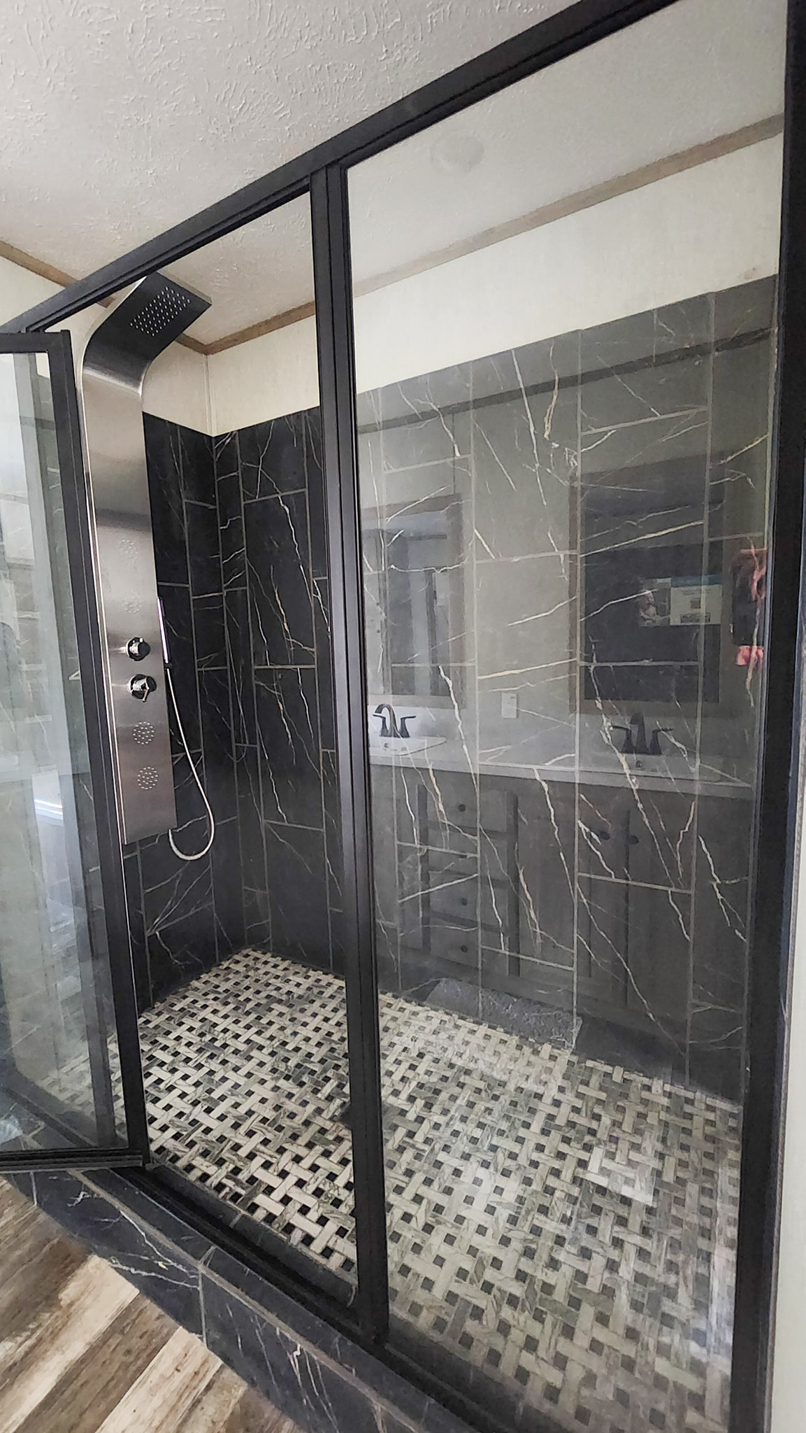 Luxurious glass shower with a sleek black frame, marble-patterned tiles, a modern shower panel, and a mosaic floor, reflecting a stylish vanity.