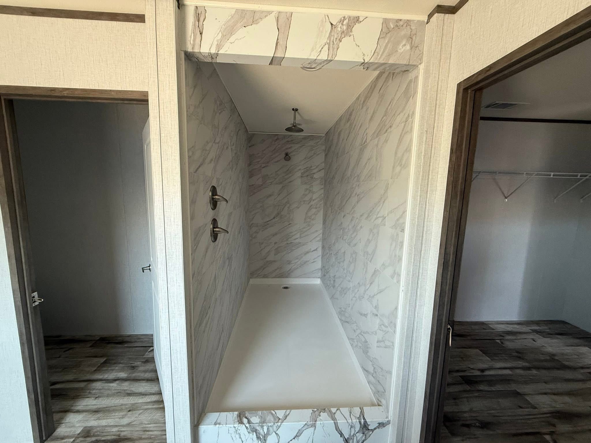 Narrow marble-tiled shower between two doorways, featuring dual shower heads on the left and a rain shower head above, evoking a modern minimalist style.