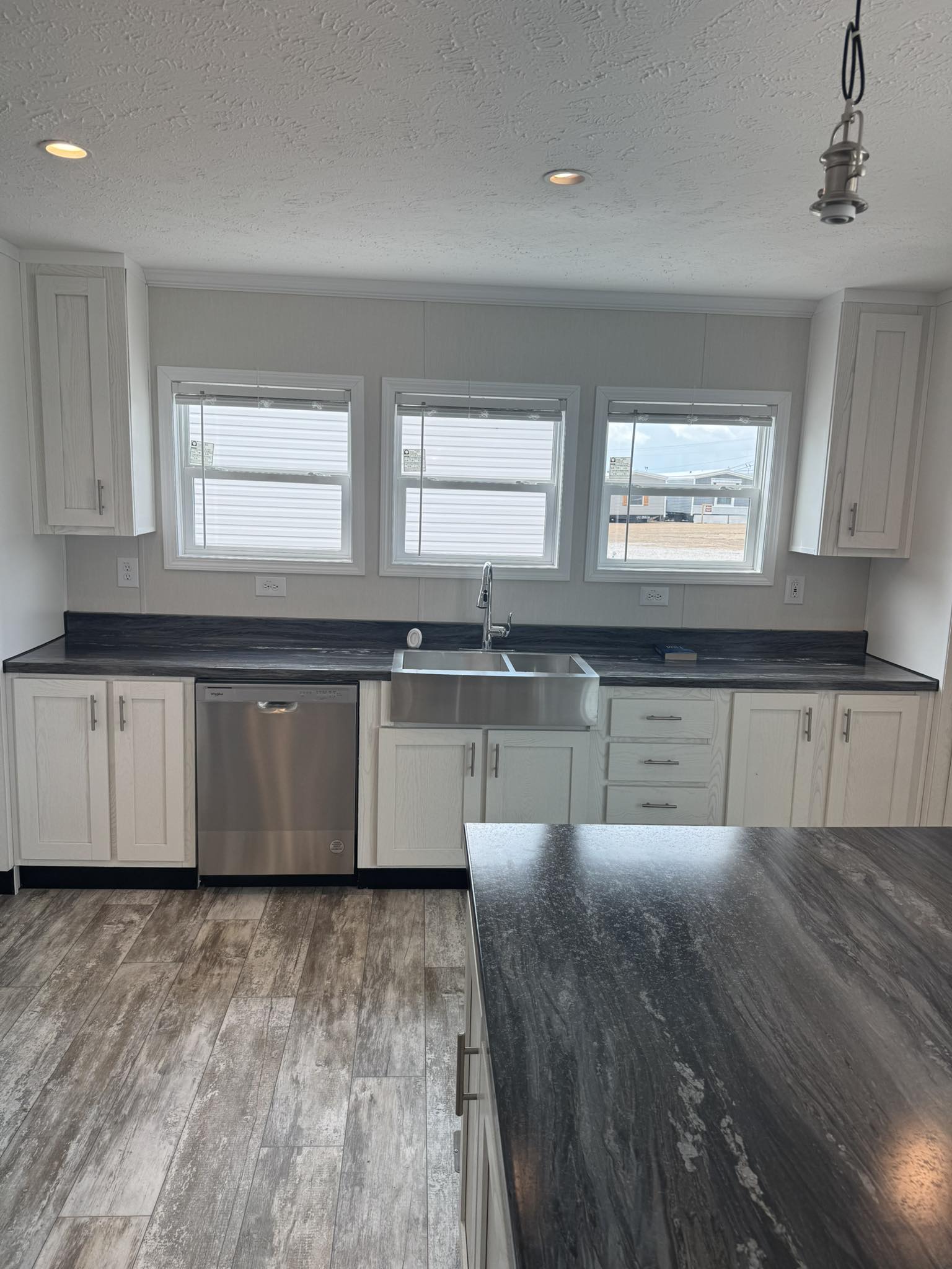 Modern kitchen with gray wood flooring, black countertops, and white cabinetry. Three windows above a stainless steel sink and dishwasher. Bright, clean ambiance.