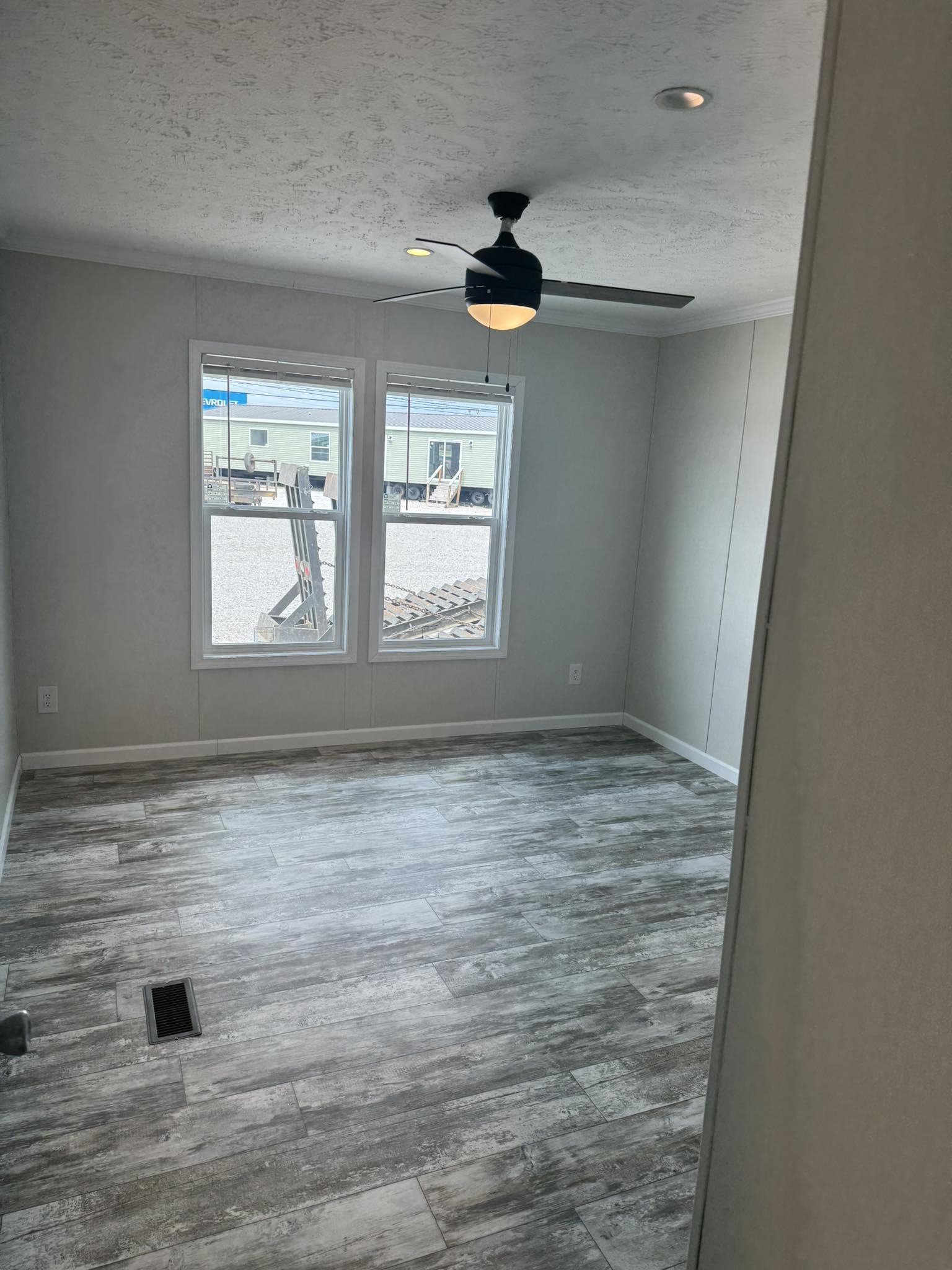 Empty room with gray wood-like flooring, white walls, and a ceiling fan with light. Two windows show a view of mobile homes under a bright sky.