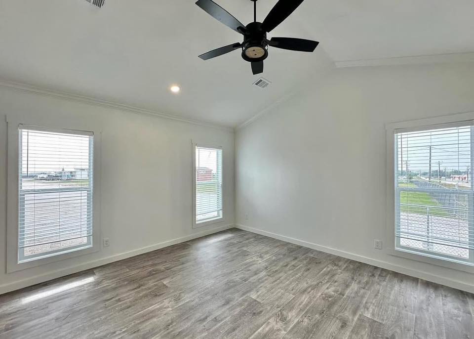 Spacious empty room with light gray hardwood floors, three tall windows on white walls, and a ceiling fan. Bright and airy atmosphere.