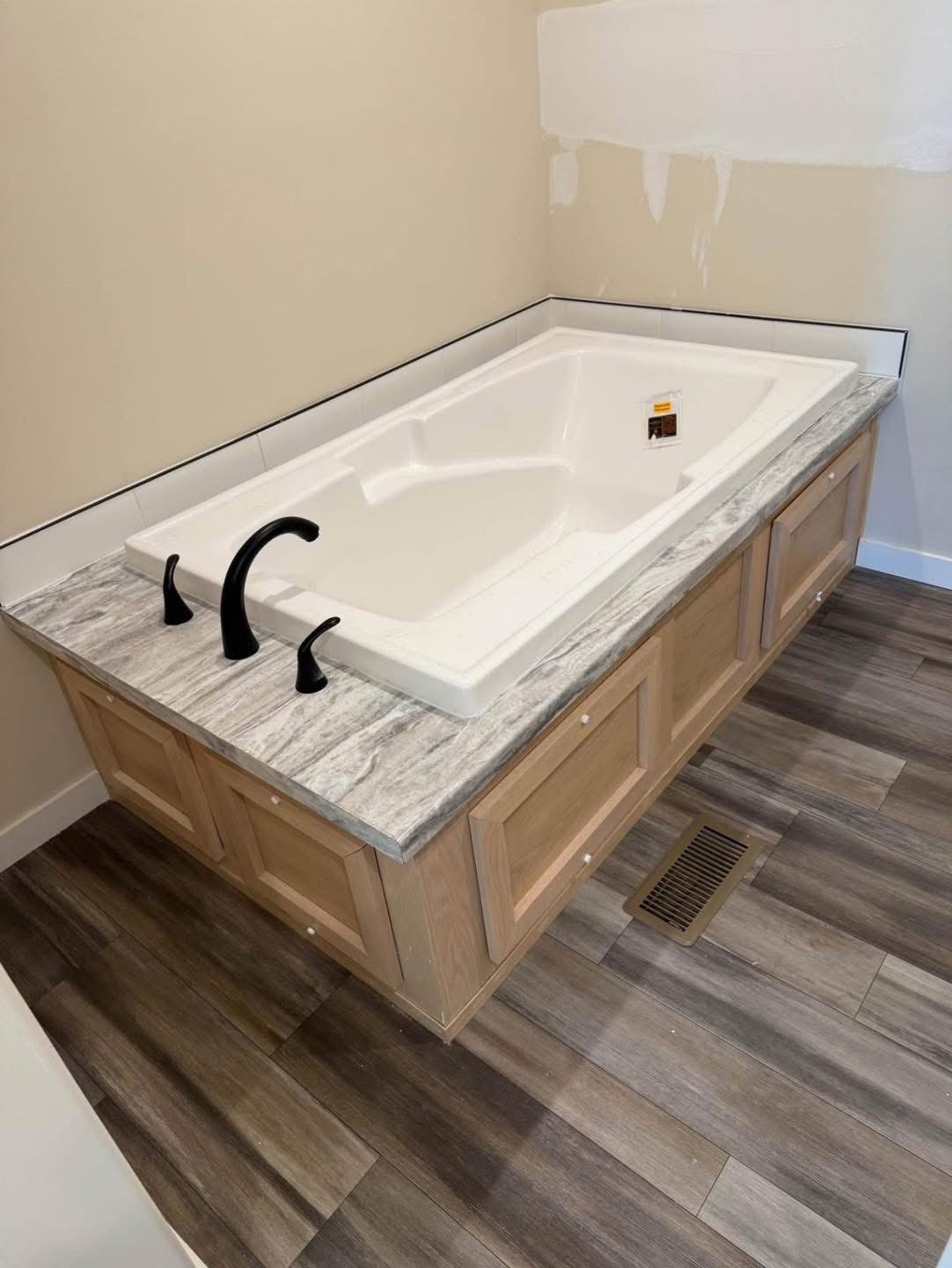 A large, white bath with a gray marble surround, black faucets, and wood paneling is set on a wood-tiled floor in a partially unfinished room.